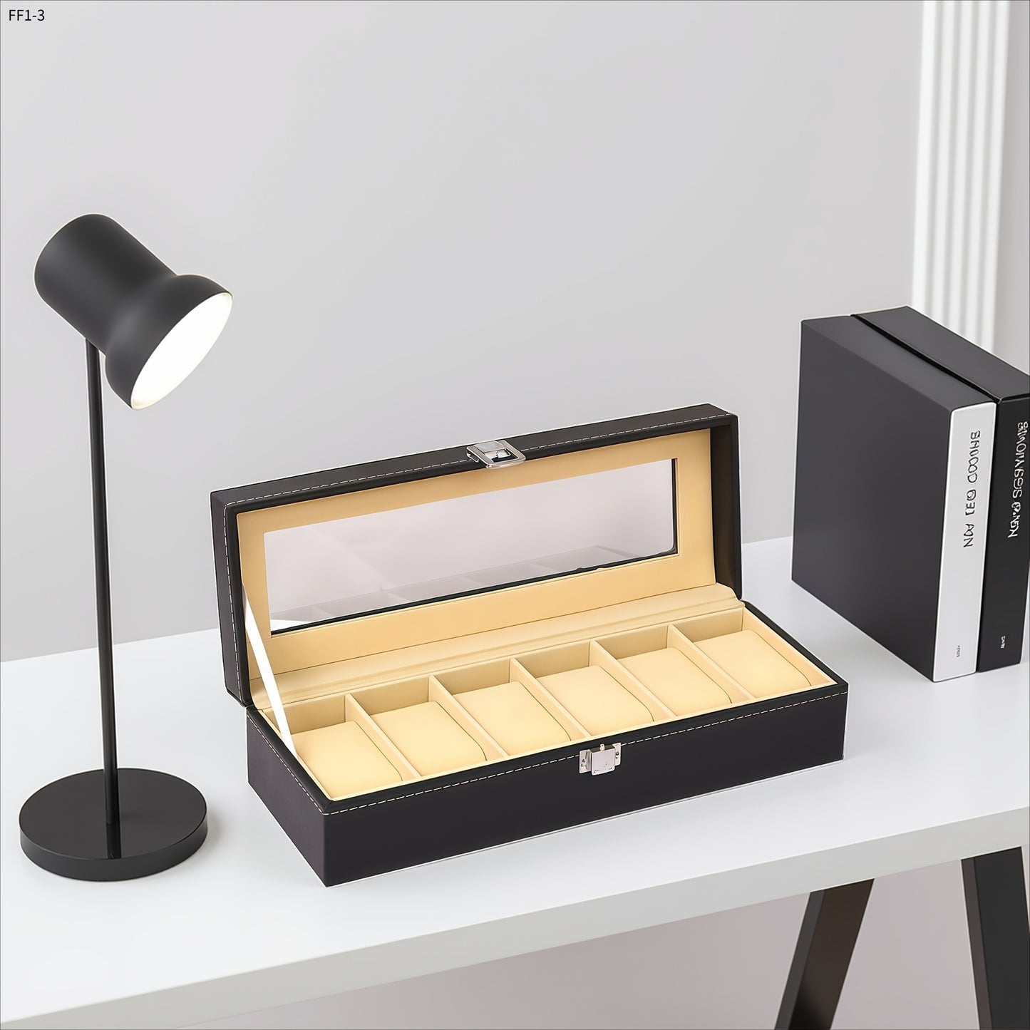 Black jewelry box with glass lid on a white surface next to a black lamp and book.