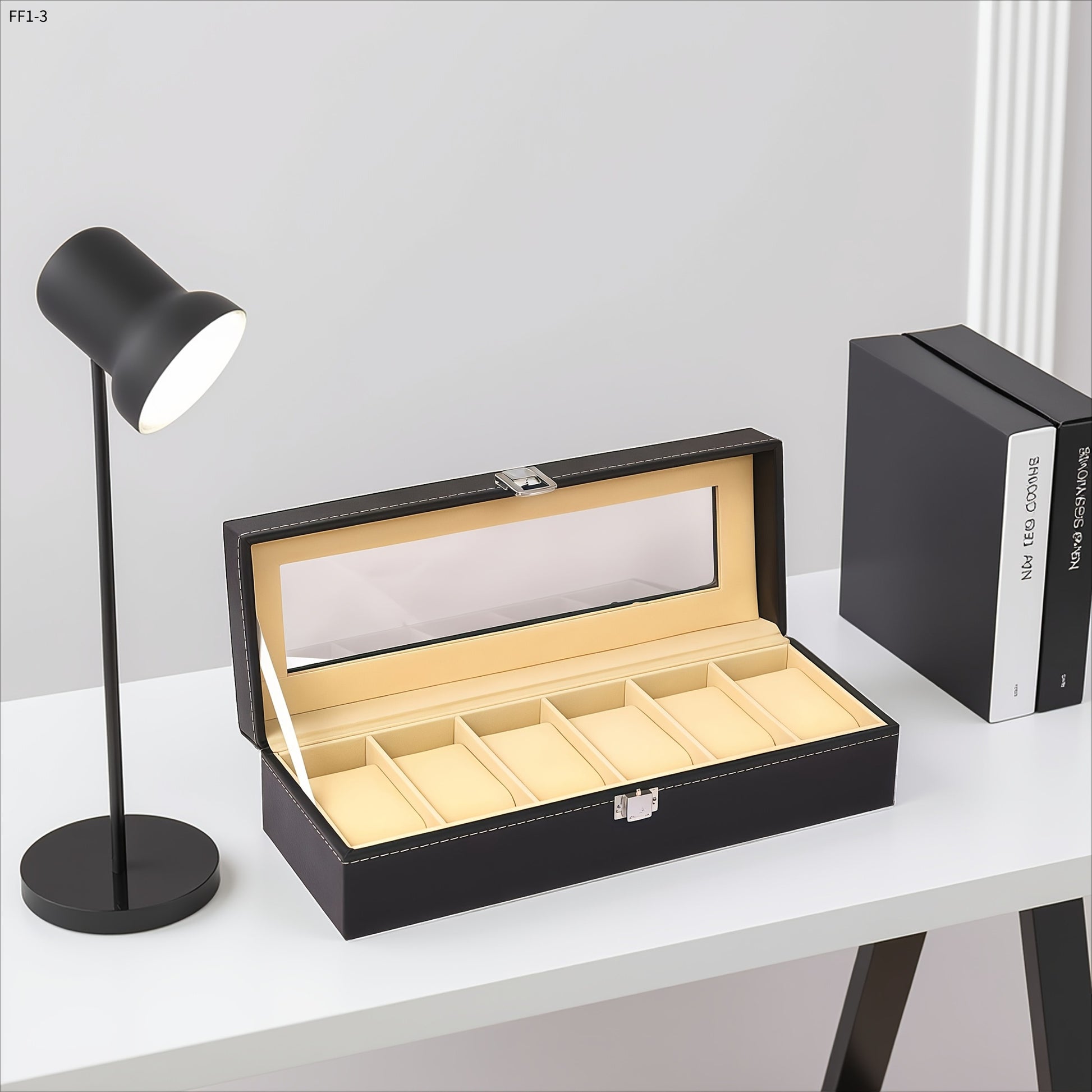Black jewelry box with glass lid on a white surface next to a black lamp and book.