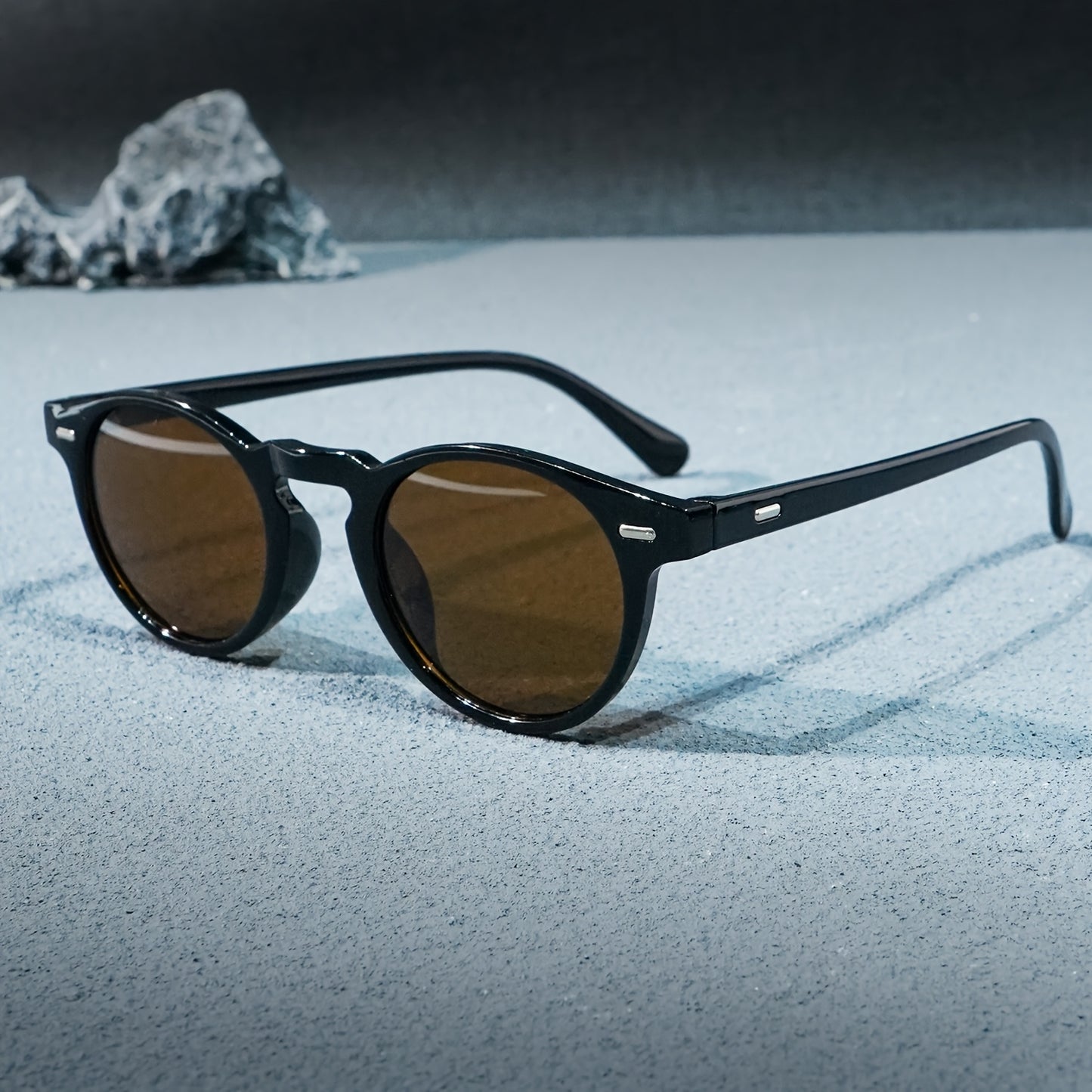 Black sunglasses with brown lenses on a sandy surface with rocks in the background