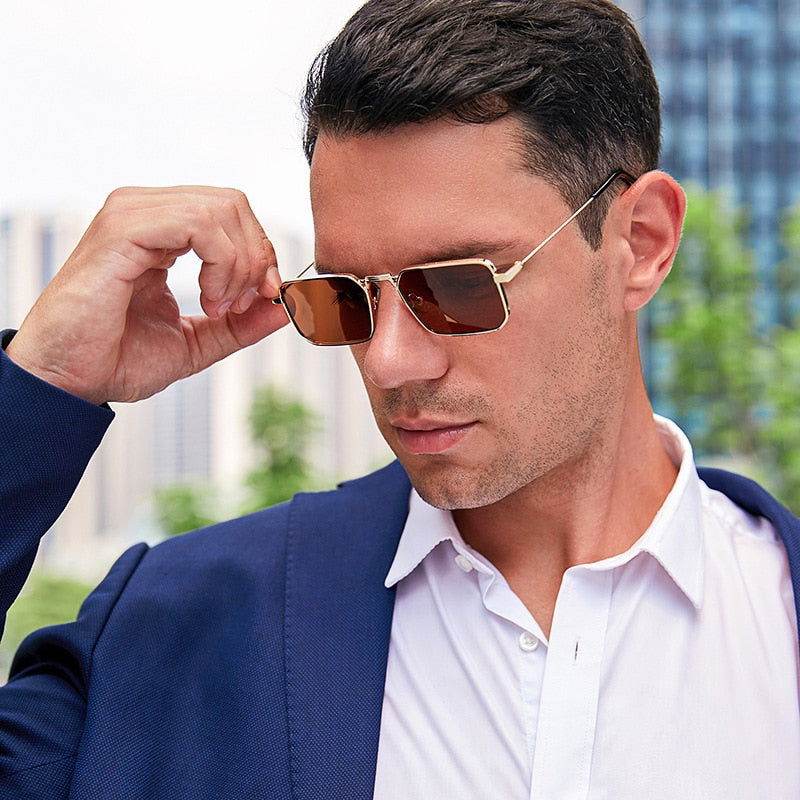Man in navy blazer wearing gold frame square retro sunglasses with brown lenses in urban outdoor setting